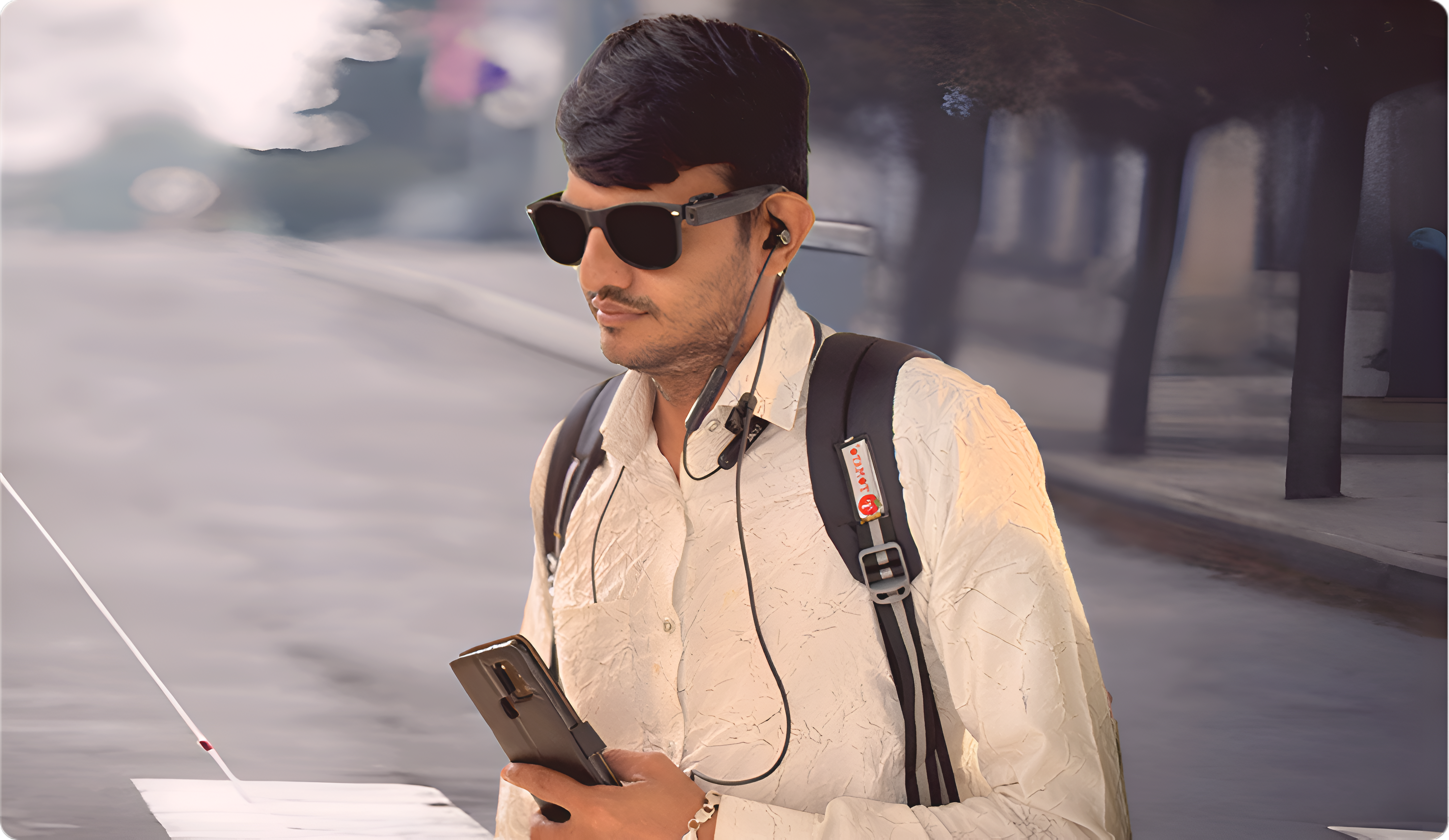 A young man on a city pavement in a white shirt and backpack, wearing dark glasses with a small camera module clipped to the right temple, an earphone cable running down to his collar, a phone in one hand and a white cane in the other.